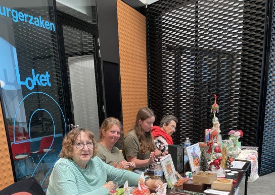 Vier vrouwen zitten achter een tafel met handgemaakte kerstdecoraties en kaarten in een moderne hal. (vergroot foto)