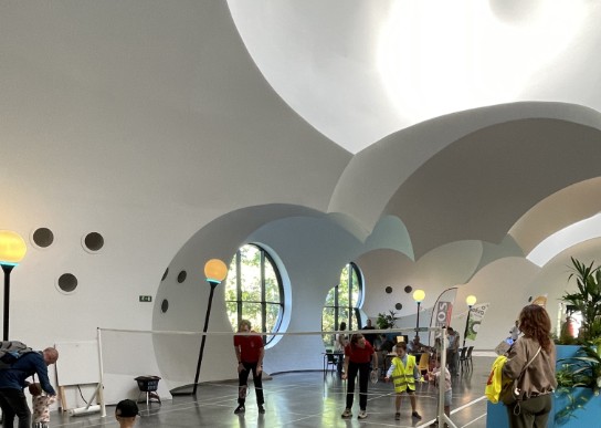 Kinderen spelen badminton in een moderne, ronde zaal met grote ramen en gebogen plafonds. (vergroot foto)