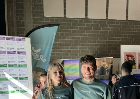 Twee mensen in groene truien met tekst "Give a Day" poseren bij een informatiestand in een hal met bakstenen muur. (vergroot foto)