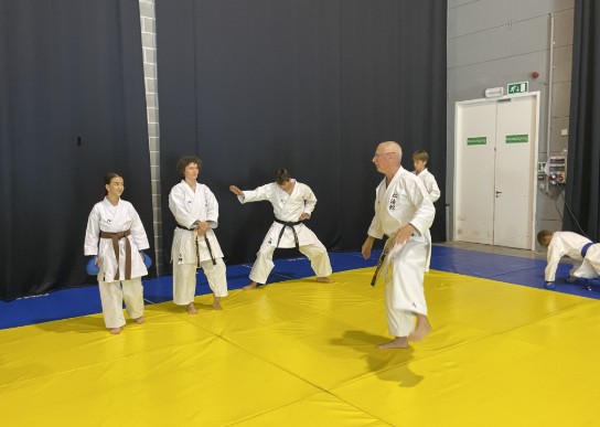 Groep mensen in witte karatepakken oefent op gele mat in dojo met zwarte wand en nooduitgang. (vergroot foto)