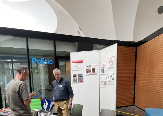 Twee mannen bij een tafel met boeken en folders in een hal met een glazen deur met het woord 'Politie'. (vergroot foto)