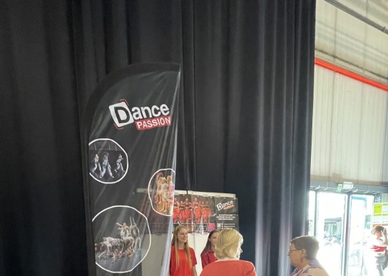 Twee vrouwen praten met twee meisjes bij een Dance Passion stand met banners en flyers in een hal. (vergroot foto)