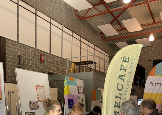 Vrouw in gele schort serveert eten aan bezoekers bij een kraam in een hal met banners en flyers. (vergroot foto)