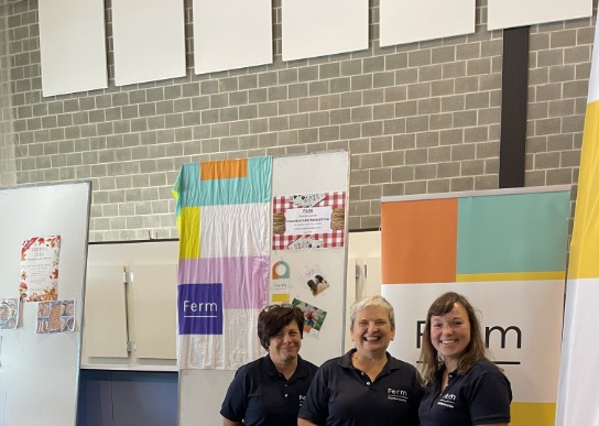 Drie vrouwen in donkerblauwe Ferm-polo's staan achter een tafel met boeken en promotiemateriaal in een hal. (vergroot foto)