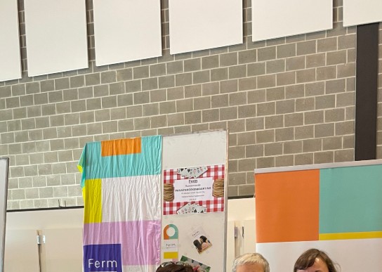 Drie vrouwen in Ferm-polo's staan achter een tafel met brochures, boeken en een laptop bij een informatiestand. (vergroot foto)