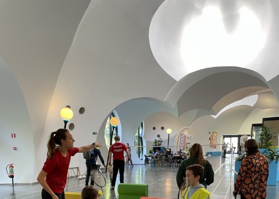 Binnen in een sporthal leren kinderen badminton van een instructrice met een racket en ballen. (vergroot foto)