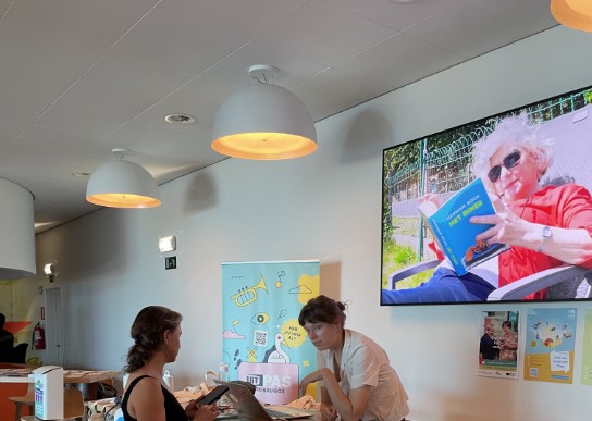 Twee vrouwen praten aan een hoge tafel in een lichte ruimte met posters en een groot scherm aan de muur. (vergroot foto)