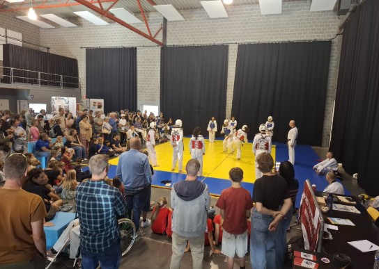 Publiek kijkt naar een taekwondo-demonstratie in een sporthal met gele en blauwe matten. (vergroot foto)