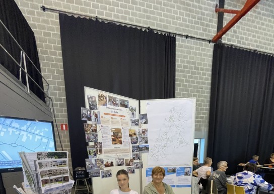 Twee vrouwen zitten achter een informatiestand met folders en een rad van fortuin in een zaal. (vergroot foto)