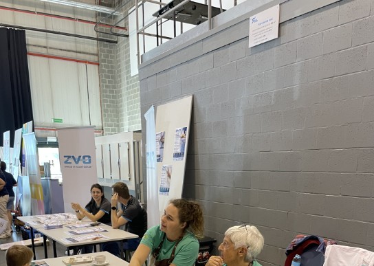 Twee vrouwen in groene shirts en schorten zitten aan een tafel met flyers, kinderen staan ervoor in een hal. (vergroot foto)