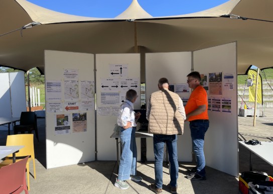 Drie mensen bekijken informatieborden onder een tent bij een evenement met stoelen en tafels eromheen. (vergroot foto)