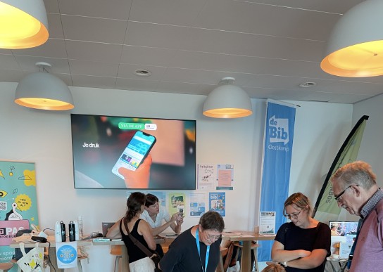 Mensen spelen bingo en bespreken informatie in een bibliotheek met borden en schermen. (vergroot foto)