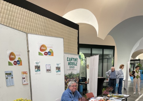 Man zit achter tafel met tijdschriften en folders bij een informatiestand in een hal met bogen. (vergroot foto)